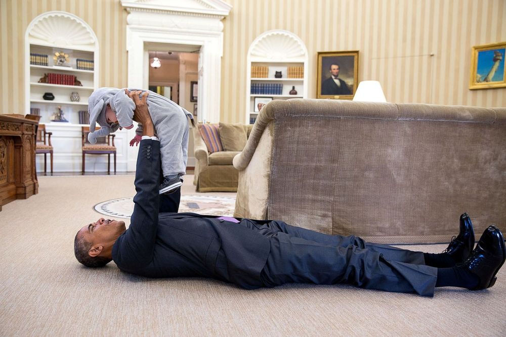 Barack Obama in the White House laying on the floor hold a child up in the air in his outstretched arms.