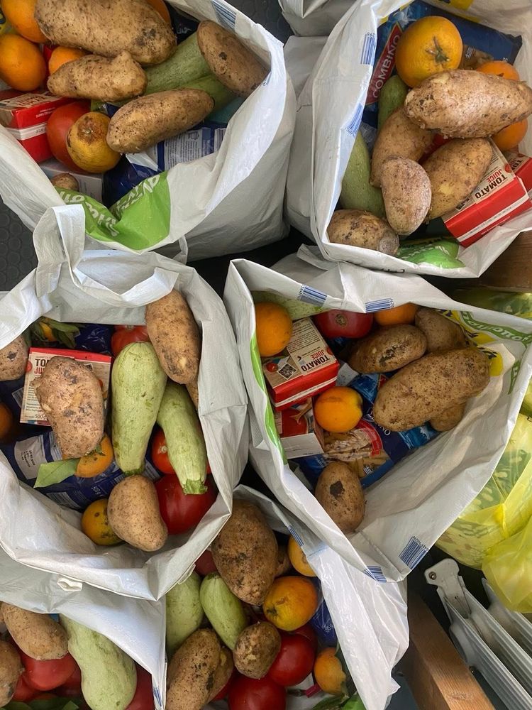 Lebensmittel-Pakete bereit für die wöchentliche Verteilung 
Man schaut von oben auf die Pakete, weiße Säcke, gefüllt mit Trockenlebensmittel wie Reis, Nudeln, Tomatenmark, Linsen, usw.
Oben auf: Kartoffel, Karotten, Zucchini , Tomaten, Zwiebeln, Orangen 