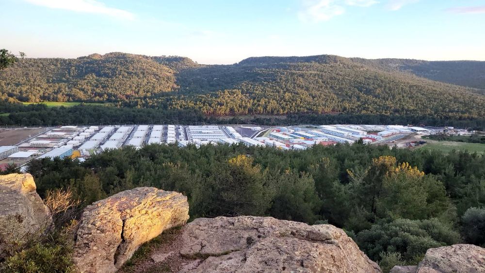 Man sieht das zukünftige Camp Vastria, mitten im Wald liegend, kilometerweit nichts, nur Wald. Im Vordergrund Steine auf einem Hügel, dahinter viele Container, alles abgesperrt durch einen hohen Zaun, oben auf Nato-Zaun. 