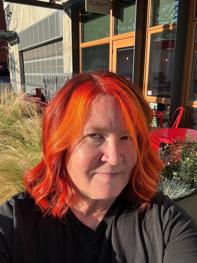 Selfie of me standing outside Paint Salon in Capitol Hill, Seattle with fresh cut and color. My hair is very red, orange and yellow, my skin is very pale and my smile is very happy