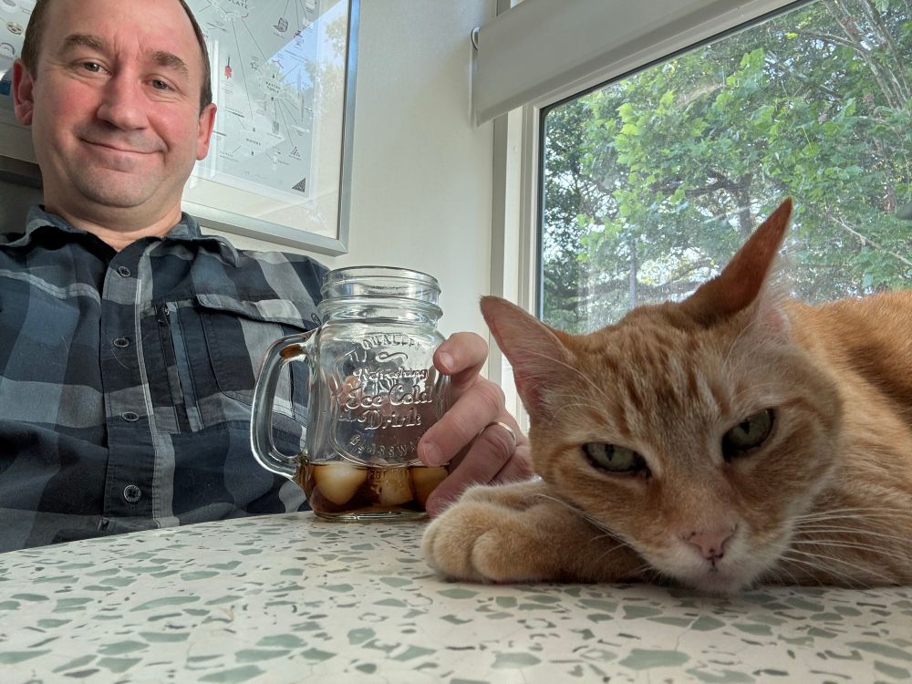 “My reluctant companion.” At left, a middle-aged nerd clutches his iced coffee defensively while catching up on his Bluesky feed. At right, Clementine, a perpetually disgruntled orange tabby, stares into the void with a mix of contempt and barely-contained fury that her human dared sit down without giving her his undivided attention. (the audacity).