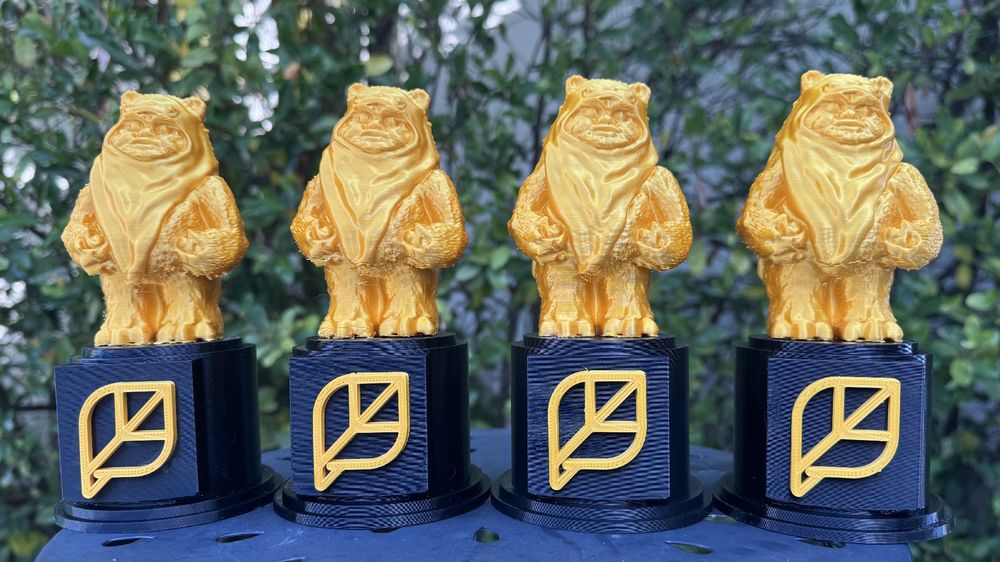 Four 3d-printed trophies with gold Ewoks on black bases with a gold company logo on the front of each.