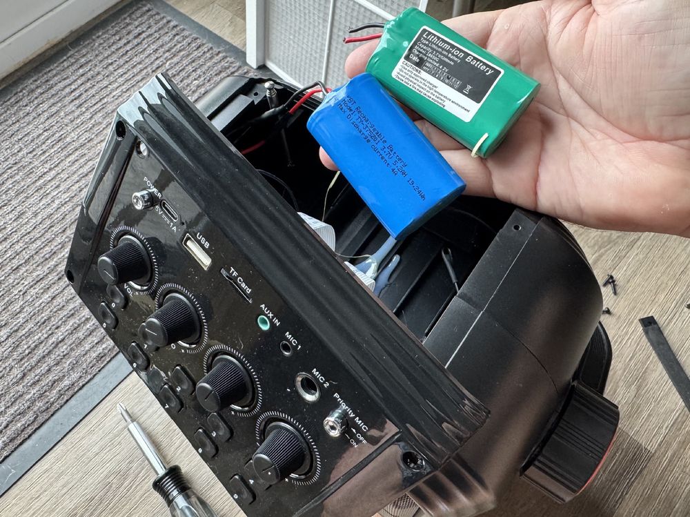 The top of a portable speaker is removed to show the battery compartment. My hand is holding both the dead battery and the new one already wired in.