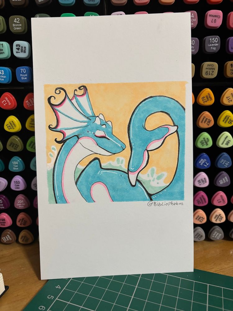 A drawing of a sea dragon, looking relaxed. They are a soft blue with pink and white markings. Behind the drawing is a collection of marker pens.
