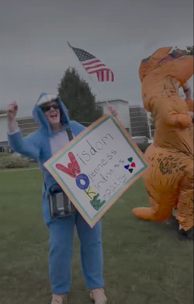Still from the video of the Gurnee, Illinois, #NoKings protest with a person dressed in a Tyrannosaurus rex costume and another holding a sign spelled WOKE with the words Wisdom, Openness, Kindness, Empathy.