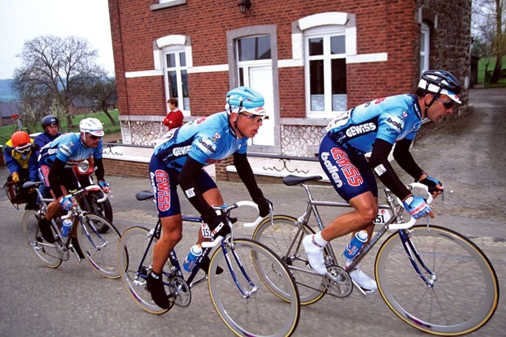 Three cyclists of the Gewiss Ballan racing team riding the 1994 La Flèche Wallone race in Belgium. Riding Italian De Rosa bicycles and blue cycling kit, they’re all out of the saddle as they take on a Belgian hill on their way to the finish as a race winning trio.