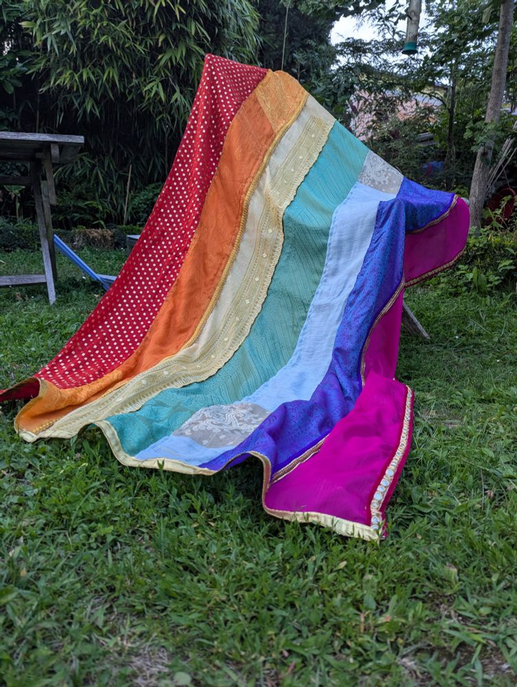 Rainbow pride flag out of Indian style scarves with gold patterns in several stripes, draped over a chair in a garden