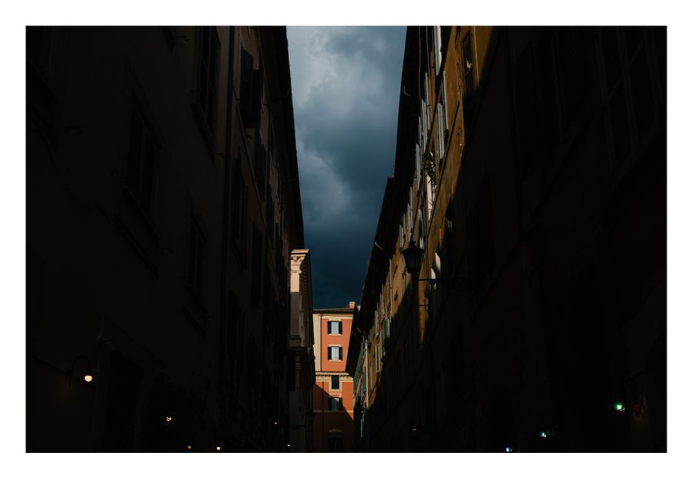 A view down a street. The buildings on either side of the street are in shadow. An orange building is illuminated at the end of the street. A dark stormy sky is above. 