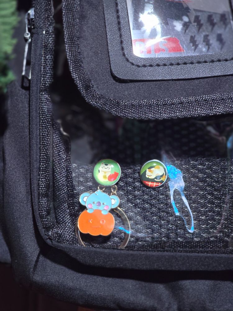A picture of the front pocket of a black ita bag, there are two small pins of cats and a charm of bt21 baby koya on top of a pumpkin secured by one of the cat pins.