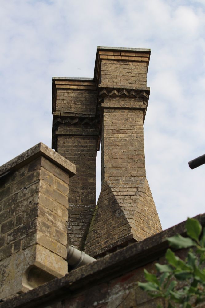Two ornamented brick chimneys behind a lower level brick parapet. Green leaves in the bottom right hand corner of the photo, a cloudy blue sky behind the brickwork.