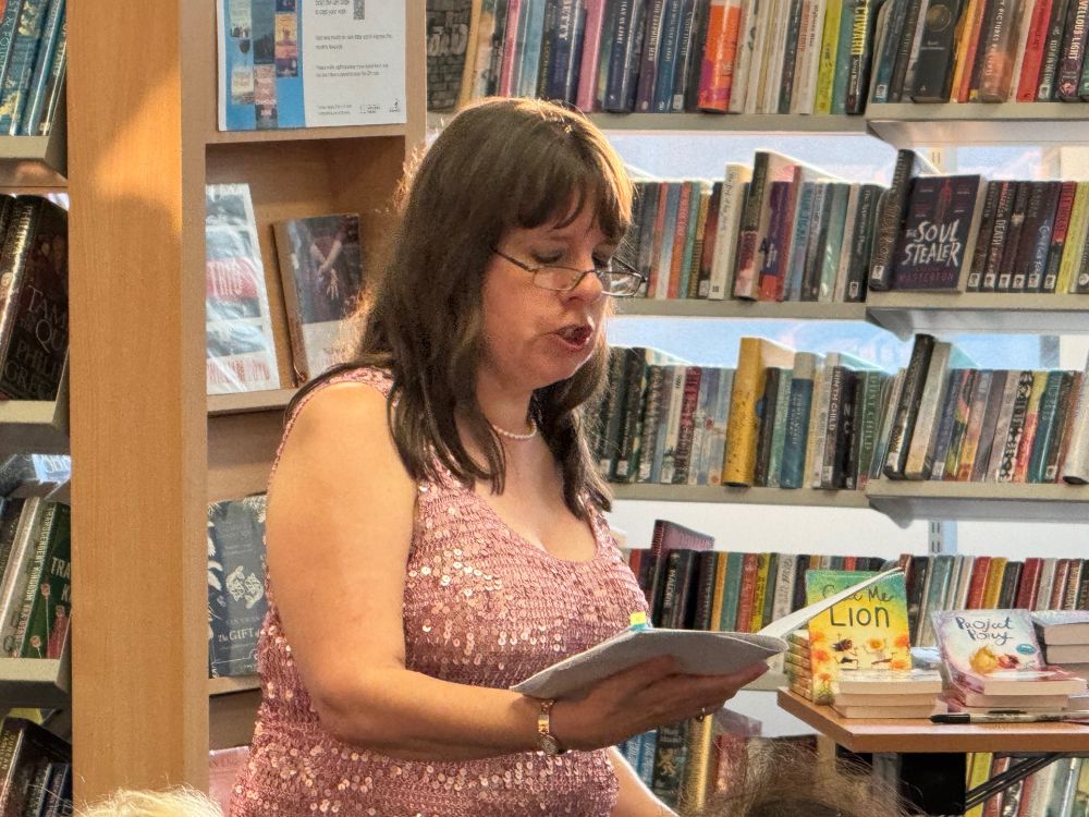 J.S. Watts reading from Cats and Other Myths. She is wearing a pink, sparkly top. Behind her are books from Hitchin Library