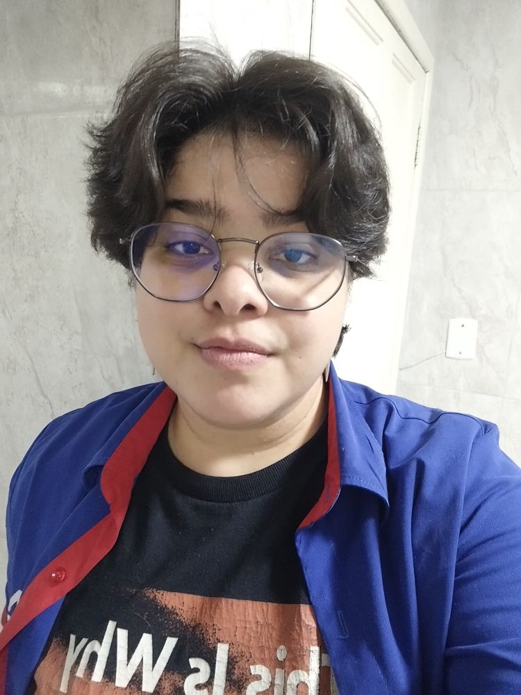 Pessoa não binária branca de cabelos castanhos.
Veste uma camiseta preta, escrita This is Why. E por cima, uma camisa azul de gola vermelha.