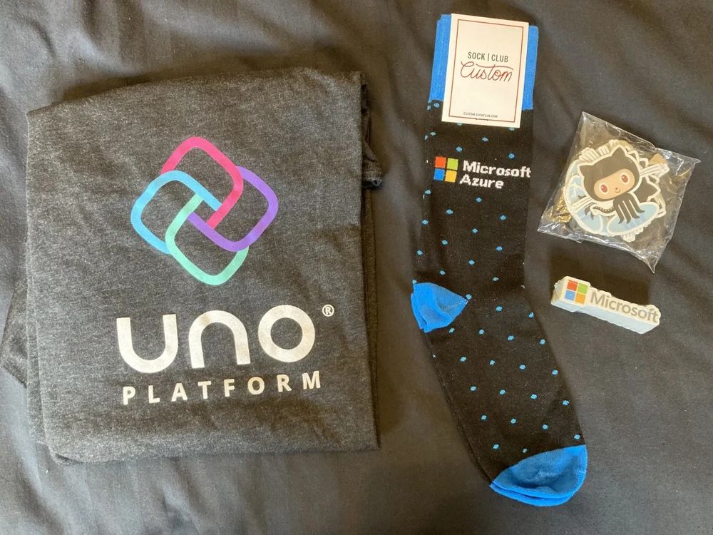 A folded t-shirt with a logo and "Uno Platform" branding, a pair of "Microsoft Azure" black socks with blue spots, and some GitHub "Mona the Octocat" and "Microsoft" logo stickers.