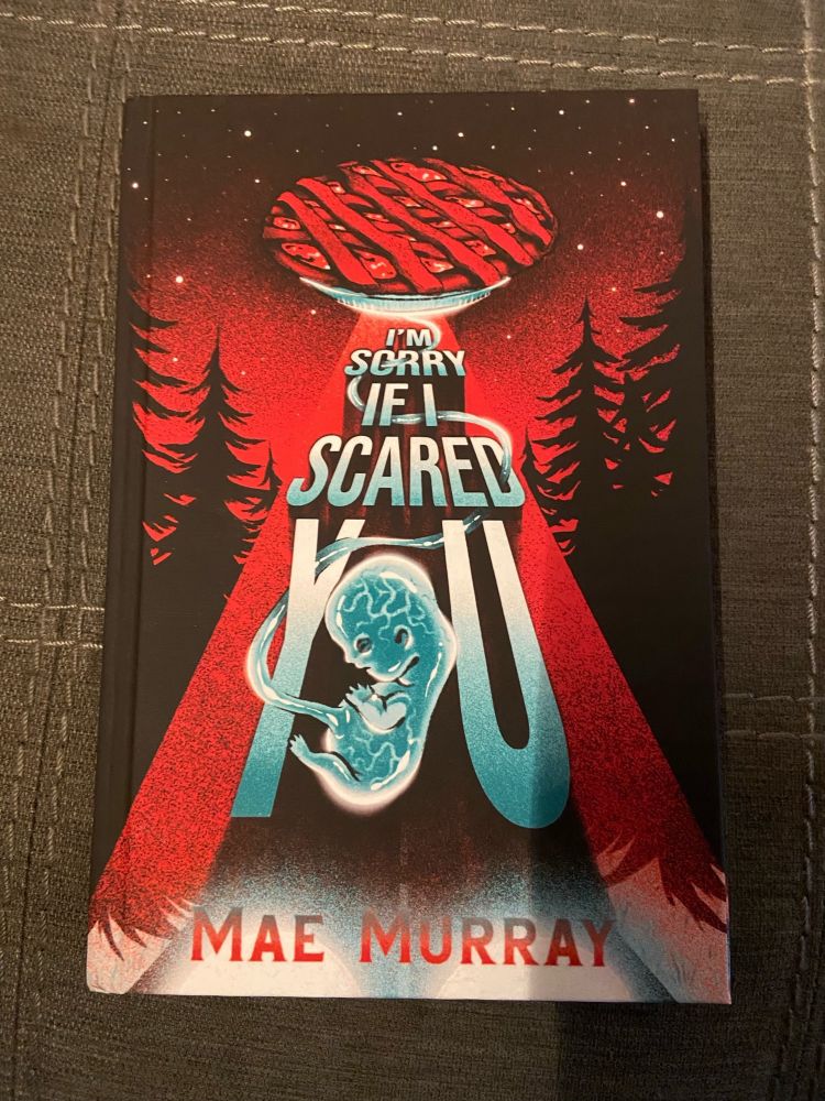 The cover of I’m Sorry If I Scared You by Mae Murray. It depicts a  pie with lattice top floating in a dark forest like a flying saucer, with an umbilical cord winding from the bottom to a fetus.