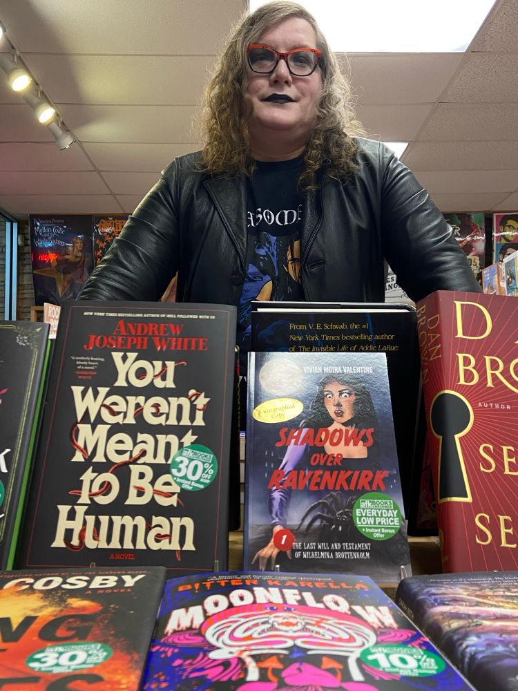 Vivian Moira Valentine, a middle-aged white trans woman, posing behind a display of books including hers, Shadows Over Ravenkirk 1. You Weren't Meant To Be Human by Andrew Joseph White stands to the left, while Moonflow by Bitter Karella is in the foreground.

Vivian is wearing a black leather trench coat and looks frankly awesome.