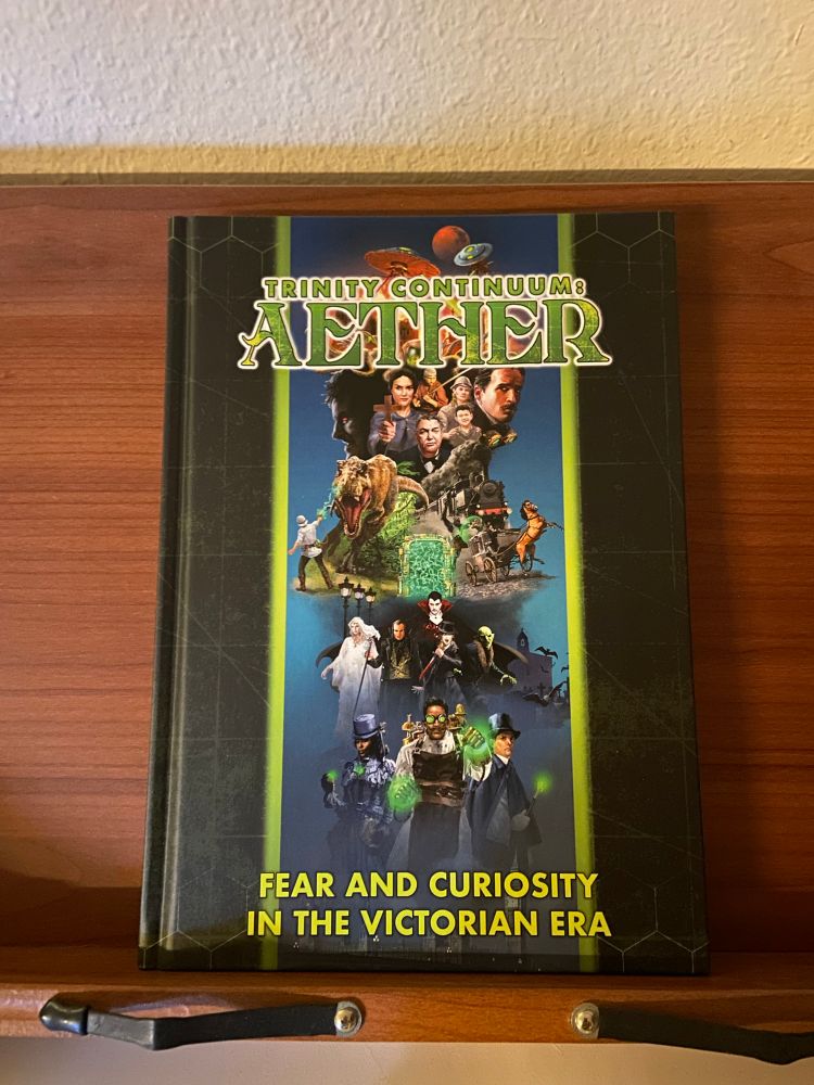 A copy of Trinity Continuum: Aether propped up on a wooden bookstand. The cover depicts a vertical collage of Victorian adventures, monsters from 1800s fiction, and mad science with an eerie green glow. The tag line at the bottom is Fear And Curiosity In The Victorian Era.