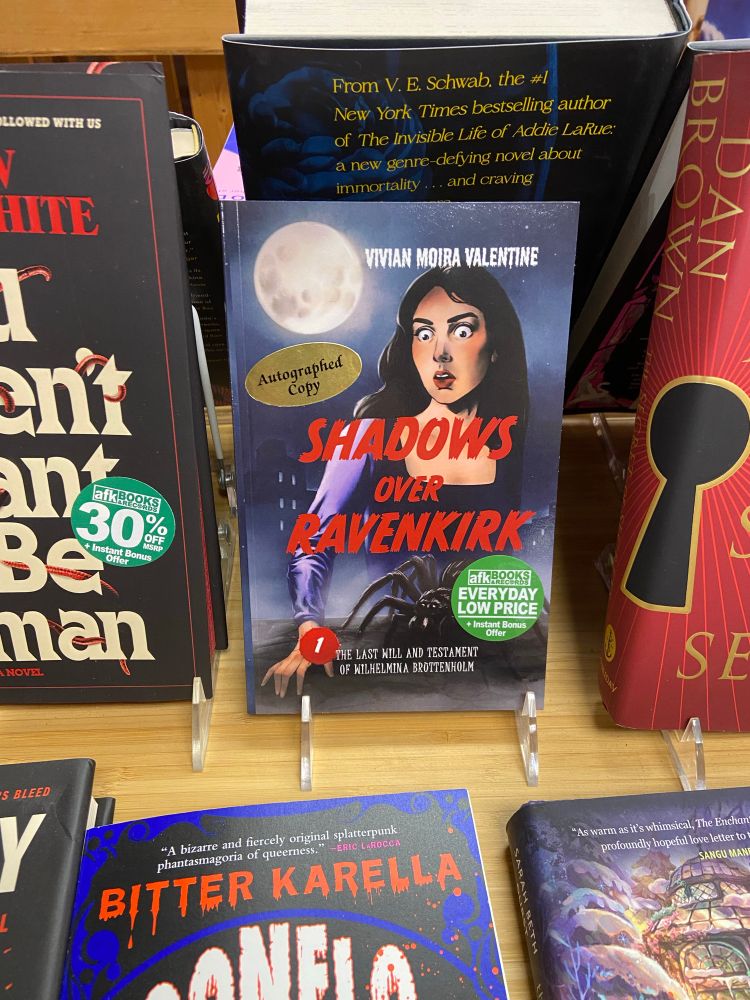 A paperback copy of Shadows Over Ravenkirk 1 displayed on a table amid other books. The cover depicts a goth woman in a black and purple dress looking on in shock as a large black tarantula crawls across a stone shelf. Behind her, mist rises, obscuring the houses in the background. A full moon hangs just above her head.

Illustrated by Lauren Walke.

Text reads: 
Top - Vivian Moira Valentine
Middle - Shadows Over Ravenkirk
Bottom - 1: The Last Will And Testament Of Wilhelmina Bruttenholm.