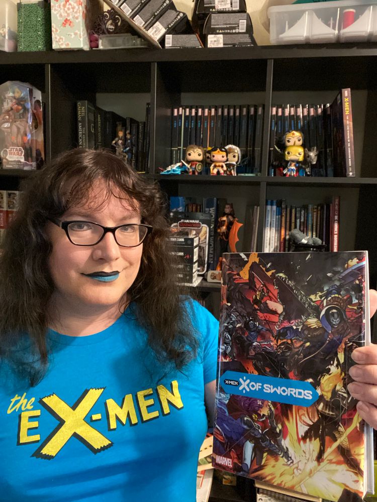 A photo of Vivian holding the massive X of Swords omnibus while standing in front of a bookshelf full of X-Men collections. She is wearing a blue shirt that says "the eX-Men" in the style of the classic logo in gold letters.