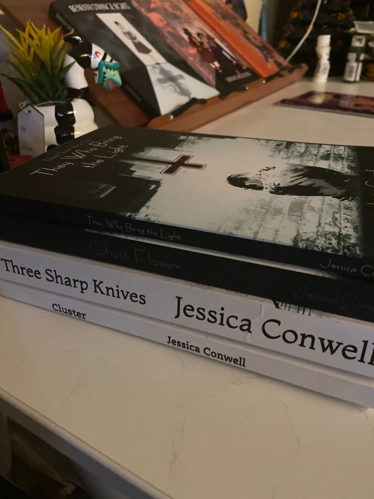 Four books by Abra Conwell, writing as Jessica Conwell - Cluster, Three Sharp Knives, Ghost Flower, and Ghost Flower Verse Two: They Who Bring The Light - sitting on the edge of my computer desk.