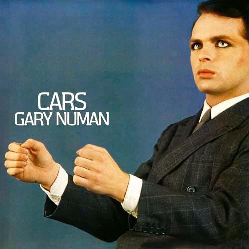 Cover of Gary Numan’s single, “Cars.” Numan is in a suit against a blue background, looking up and to the side. His hands are posed as if gripping a steering wheel, but there is no steering wheel.