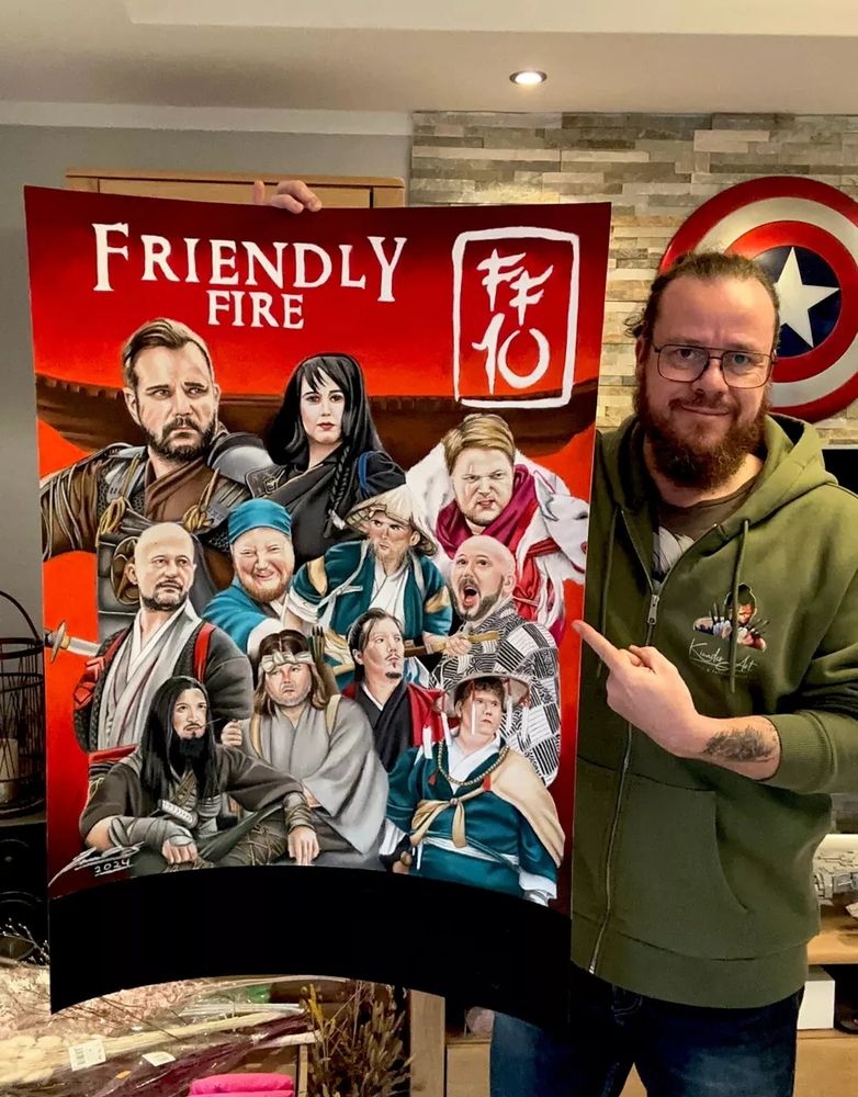 Sven Kinader hält sein Artwork zu Friendly Fire 10 in der Hand und zeigt mit dem Finger darauf. Es sind alle Talents von Friendly Fire zu sehen in ihrem jeweiligen Kostümen, gezeichnet von Kinader. 