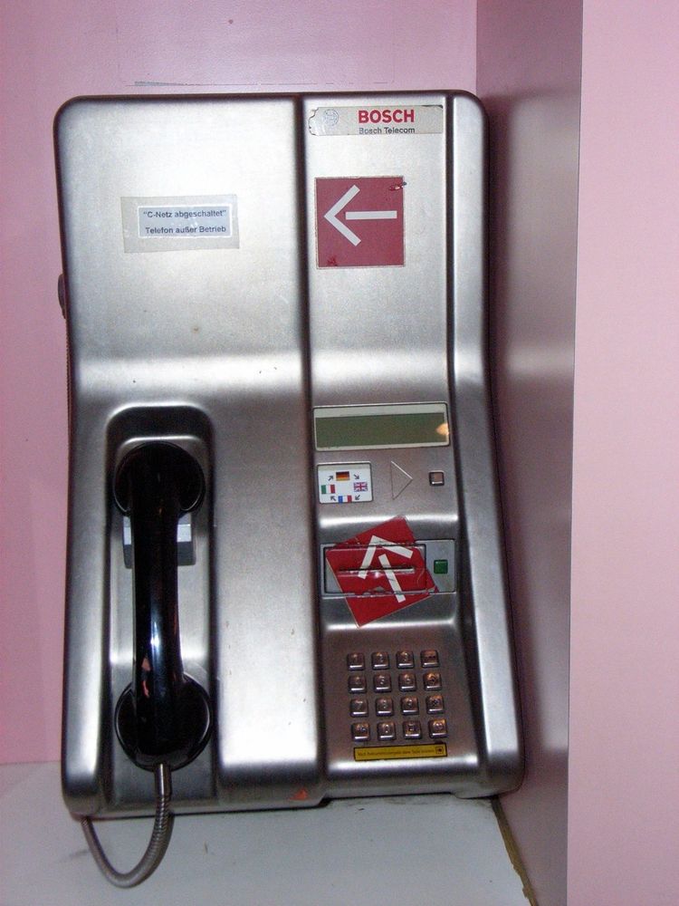 A silver public telephone booth with a black handset, featuring various buttons, language options (Italian, English), and several stickers, including a warning that indicates the phone is out of service. The booth is positioned against a pink wall.