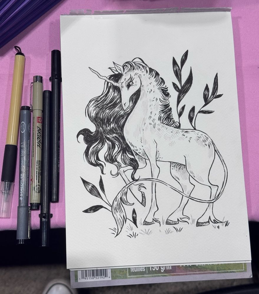 A photo of an ink drawing with some ink and brush pens to the left of the paper. The drawing is of a unicorn with a long mane, a lion’s tail and a few leaves of vegetation behind it.