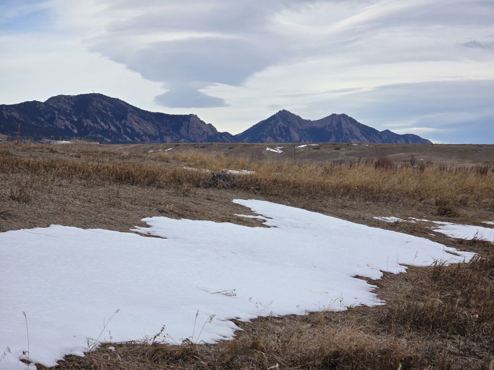 Snowdrift in foreground, dry grass meadow in middle ground, Flatirons rock formations in the distance, white and gray clouds in sky. Long Lake Regional Park in Arvada, Colorado