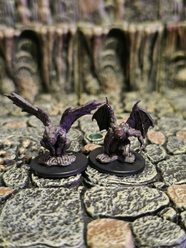 Two custom painted miniatures of gargoyles. They have stone-gray bodies with glowing red eyes. The one on the left is crouched down and hunched over, their wings extended up. The right one is crouched down and leaning forward, their right hand braces against the ground and their left hand pulled back as is preparing to scratch something.