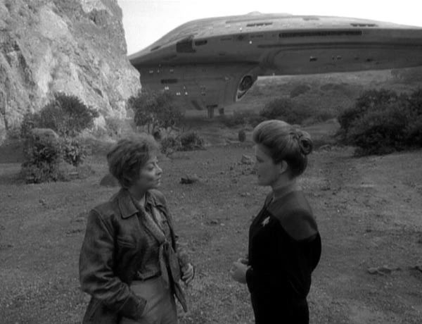 A woman in a 1930s aviator outfit amiably discusses something with another in a future military jumpsuit. In the background a spaceship is parked.