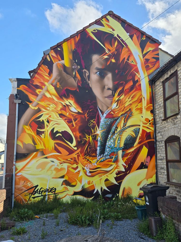 The side of a building is painted with flames, mostly obscuring the photo-realistic face of a martial artist, who has a determined expression. He is holding a yellow pole in a manner that he might either throw it like a javelin, or bring it down hard.

Bottom left hand corner is a stylised signature of the artist Jagun