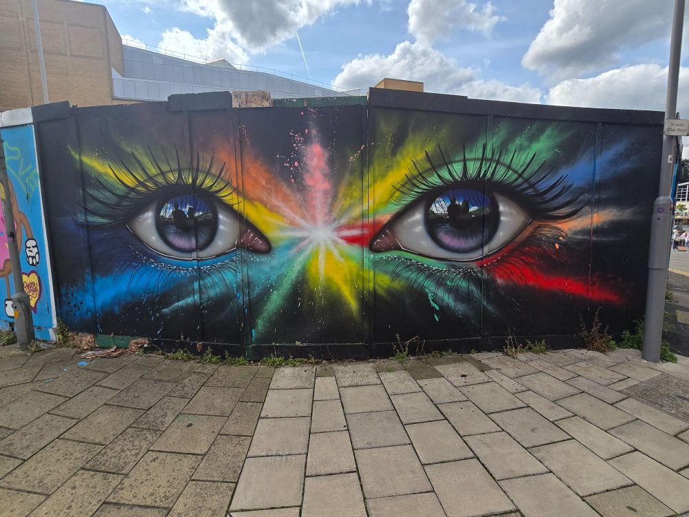 Security hoarding, with a pair of female eyes spray painted on it. There  are splashes of bright colours radiating out from the centre between the two eyes. 

At first glance, the eyes look normal, but closer inspection of the pupils show it is a reflection of two people in silhouette form, seated and facing away.