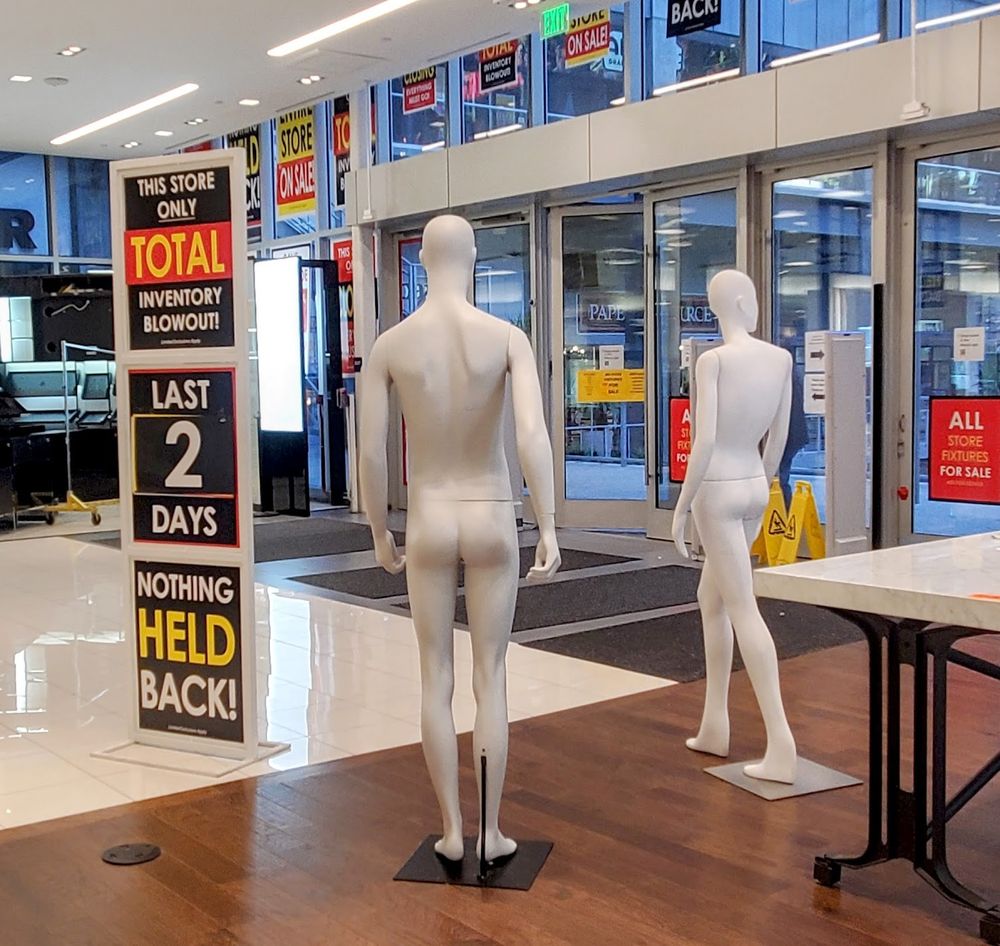 A closing department store with SALE signs all over the glass windows. Two mannequins (a man and a woman, both not dressed) stand next to a tall sign that reads "this store only, total inventory blowout, last 2 days, nothing held back"