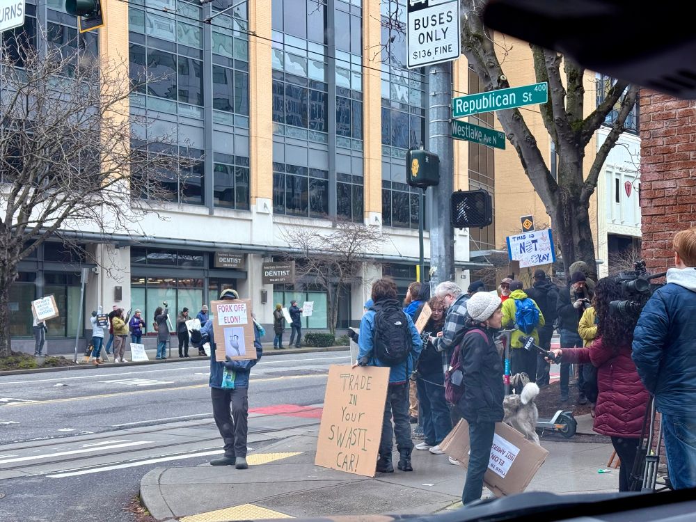 A group of 40+ people protesting Tesla outside a Tesla showroom in Seattle. They’re holding up signs saying things like “Trade in your swasti-car”