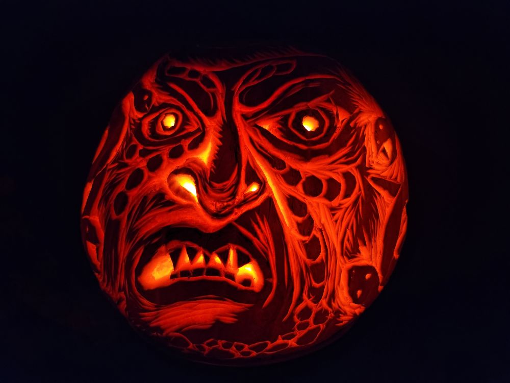 A pumpkin carved to look like a scary face with snakes for hair. The pumpkin is sitting in the dark, glowing from within