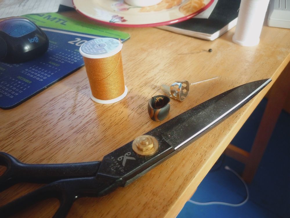 A few objects lie on a wooden desk. A pair of black sewing shears, a spool of thick yellow thread, an autoharp pick, a tiny needle, and a small rubber ring thimble.