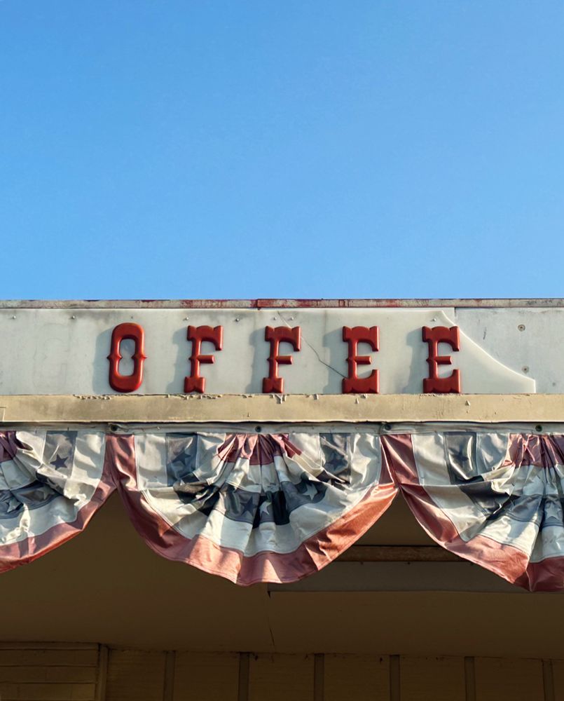 Photo: Vintage red sign letters on the side of a building spelling the word Coffee, but the C is missing (just a ghost of the letter remains). There is a blue sky above and red white and blue bunting below.