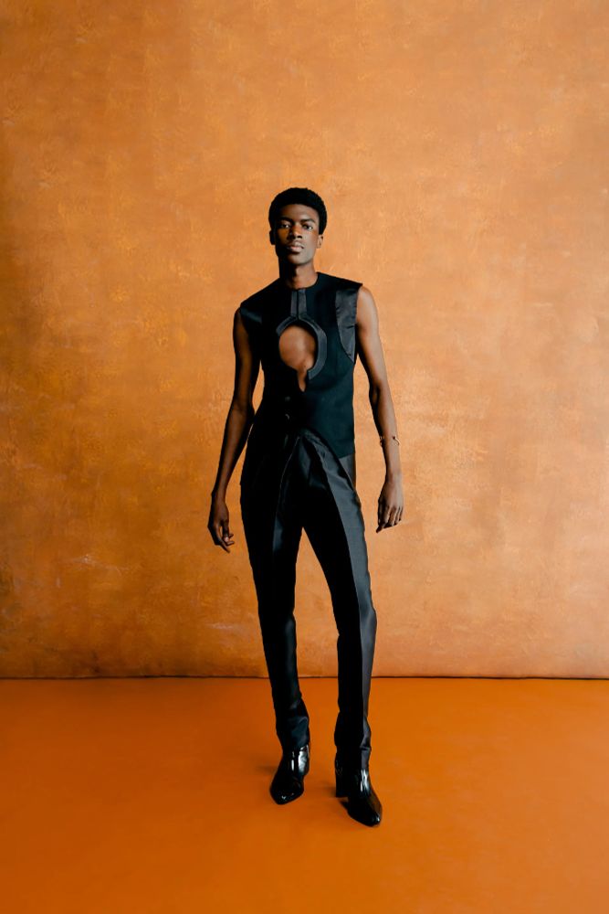 The model wears an all-black outfit. The top is a sleeveless cut-out blazer with a keyhole cutout directly in the middle of the chest. It has two buttons on the front, pinning the blazer to the side. The main body of the blazer is black crepe fabric, with black satin highlights along the arms, cutout, and hem. The model wears matching structured pants with a cummerbund style waistband. 