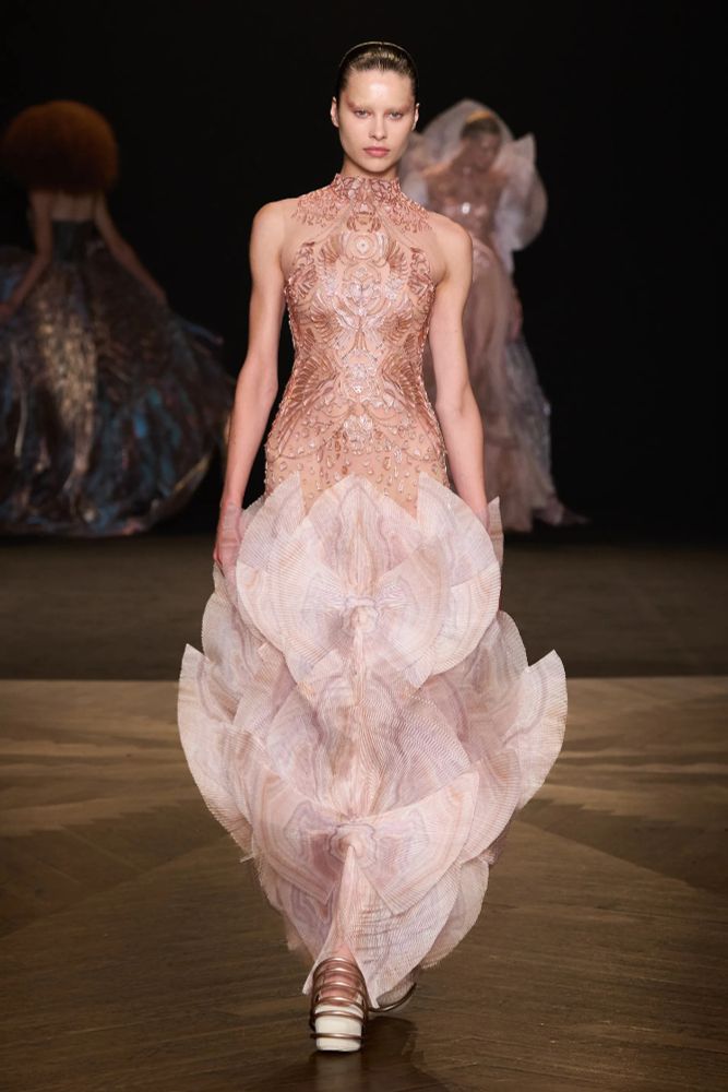 The model wears a high-neck, sleeveless "mermaid" gown in light pinks, creams, and plums. The torso of the dress is made of numerous lasercut, organic designs while the skirt is made up of dozens of fin-like panels that end at ankle length. 