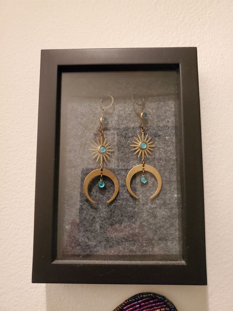 a shot of some lovely @weltschmerz.bsky.social earrings in a shadow box on my hallway wall. these are brass, with aqua-teal stones. they have lever back ear wires, then a single bezel set gem, then a sun shape with another bezel set gem in the middle, and the bottoms are crescent moon shaped, with the points of the moon toward the floor, and there is another bezel set gem in the hollow created in the center of the moon.