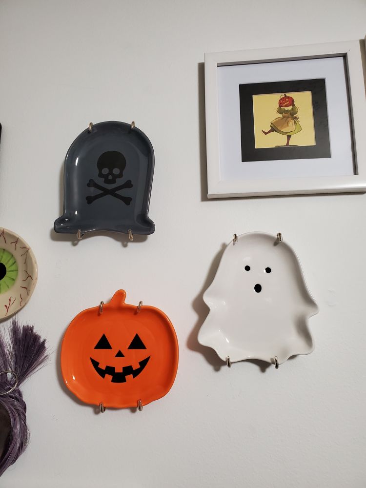 a close up of the newest plates. a grey tombstone with a black skull and crossbones, a bright orange pumpkin with a traditional happy face, and a ghostie making an :o face. i have hung them on the wall in my office, above my desk, where there was some blank space between my other plates and some framed pumpkin girl art.