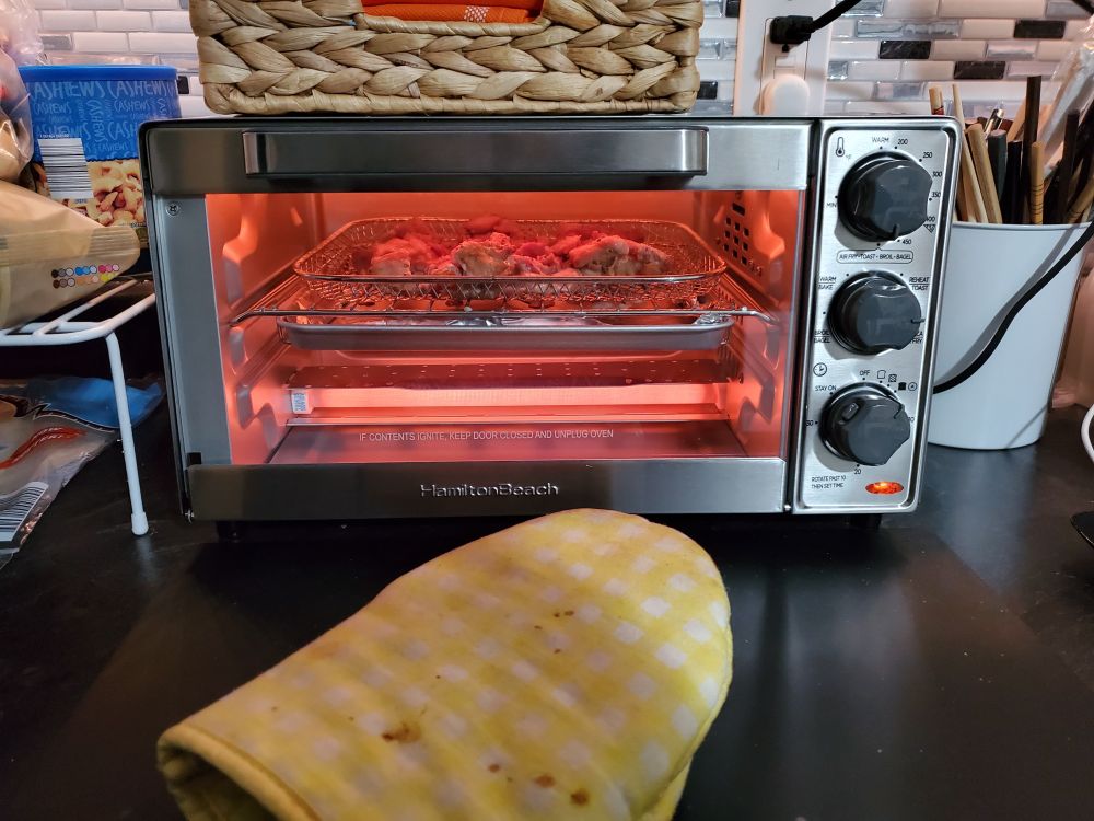 the new toaster oven, on the counter where the old one used to be. there is cubed seasoned chicken in the air fryer basket, foil on the drip tray for easy clean up, and a pot holder sitting on the counter in front. the machine is on, so everything has a red-orange glow.
