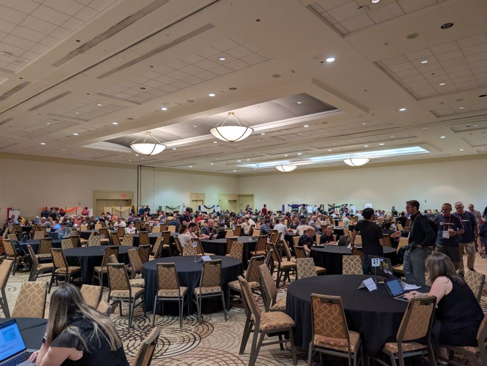Hundreds of people gathered around computers and round tables, working.