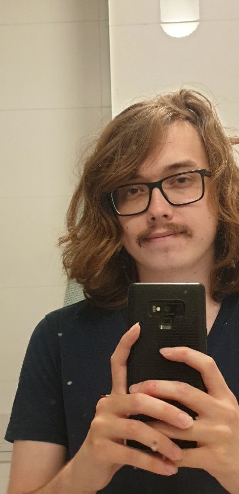 young man with long hair, square black glasses and moustache smiling