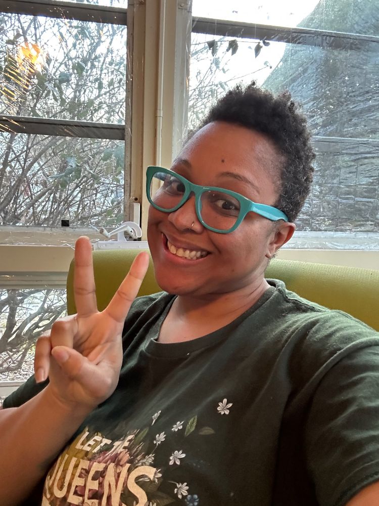 Black person with a fade hairstyle and blue glasses wearing a green t-shit sitting on a green chair. They are smiling and holding up the peace sign. 
