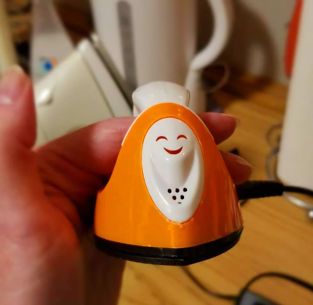 Miniature bright orange electric fabric iron suspended by the handle on two fingers so that we are looking at the front point of the iron as it would lay flat.
Where the spray and steam vents would be on a standard steam iron, this tiny iron has a decorative white plate with fake vent holes in the shape of a very smiley face.