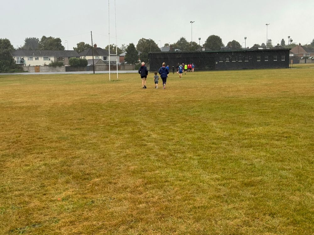 A group of runners running on a playing field. The picture shows the main pack of runners from behind. One runner is a small child 