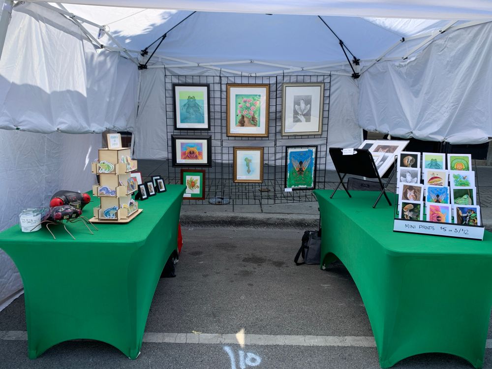 Photo of an artist tent at a fest with green tables and a lot of bug and bird related art on display including a painted plaster cicada and a cicada mask/hat