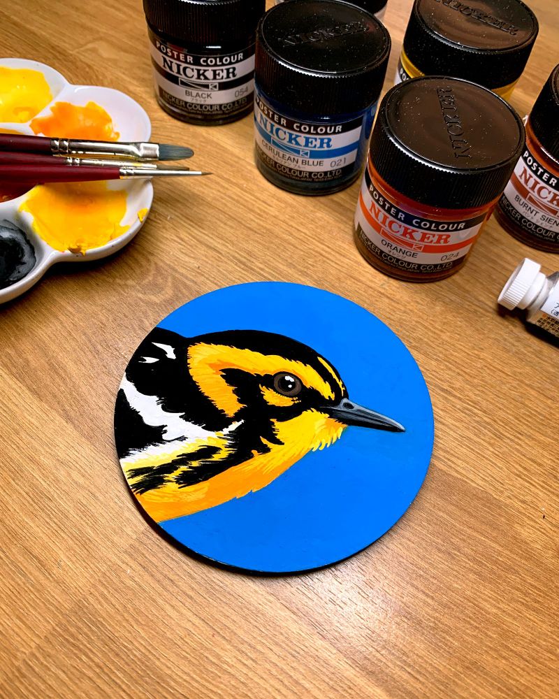 Photo of the same painting on a desk with small jars of paint and a palette with brushes also visible