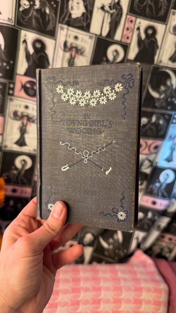 holding up a hand-bound journal with an old book cover. the book is titled "A Young Girl's Wooing" by E.P. Roe.